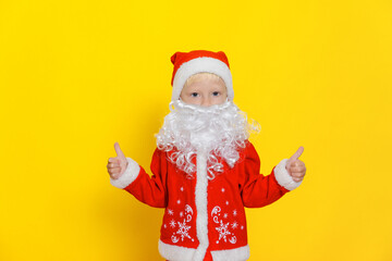 Fototapeta na wymiar Little boy in santa claus costume with white beard shows thumbs up gesture with hands, stands on yellow isolated background.