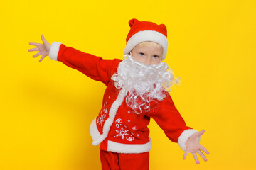 Caucasian child in New Year's Santa Claus costume spread his arms in different directions showing a gesture that he wants to hug or glad to meet you, yellow isolated background.
