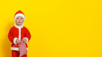 Fototapeta na wymiar A child in a red New Year's suit stands with a bag of gifts in his hands, mysteriously looks into the camera. Copy space, yellow background.