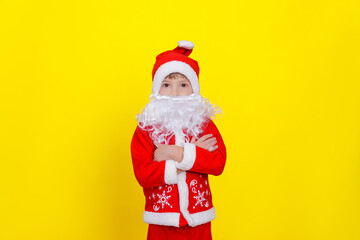 Fototapeta na wymiar A boy in a .d santaclaus suit and an artificial beard stands on a yellow background and crosses his arms on his chest, looks at the camera.