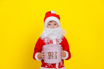 Cute little Caucasian boy in Santa Claus clothes and with an artificial beard holds a pink round gift box with the words New Year 2022 in his hands, squints and smiles, yellow studio background.