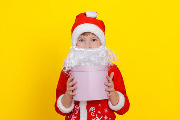 Close-up portrait of a cute child in Santa Claus clothes and with an artificial beard holding a pink round gift box in his hands, yellow studio background.