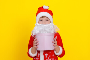 Fototapeta na wymiar Close-up portrait of a cute child in Santa Claus clothes and with an artificial beard holding a pink round gift box in his hands, yellow studio background.