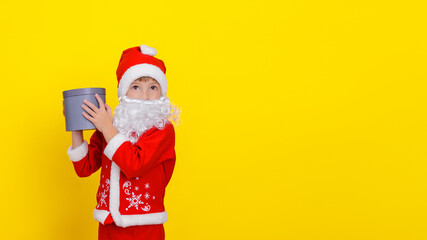 Fototapeta na wymiar Little boy in Santa Claus clothes and with an artificial beard holding a round gift box in his hands, his gaze is directed to the side, yellow, copy space.