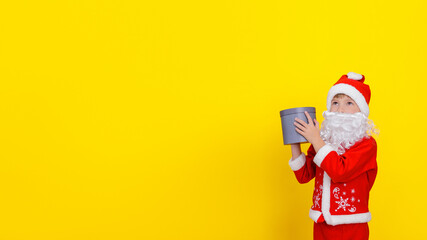 A child in Santa Claus clothes and with an artificial beard holds a round gift box in his hands, his gaze is directed to the side, yellow, copy space.