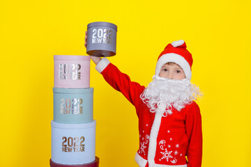 A boy dressed in the clothes of Sanat Claus picked up the topmost gift box with the inscription New Year 2022, he has a white artificial beard, stands on a yellow background.