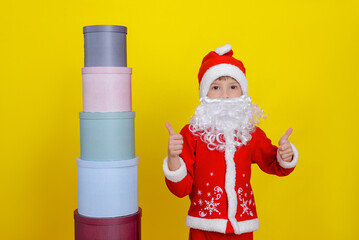 A child in Santa Claus clothes shows a thumbs up gesture, stands next to a tower of round gift boxes.