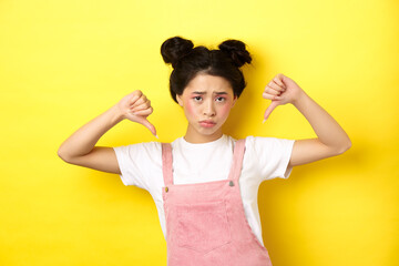 Summer lifestyle concept. Sad and gloomy asian woman with makeup, pointing fingers down and frowning upset, disappointed with unfair situation, yellow background