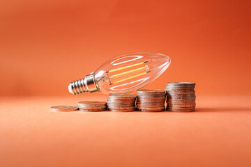 led light bulb on top of coins in the form of an ascending graph, symbolizing the rising cost of...