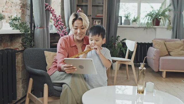 Medium slowmo shot of happy young Asian woman and her cute 3-year-old son having video chat with family or friends on digital tablet at cozy apartment. Boy blowing party horn