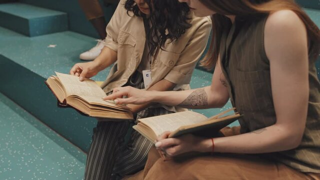 Midsection slowmo shot of two unrecognizable female university students sitting on turquoise stairs indoors reading books and discussing content