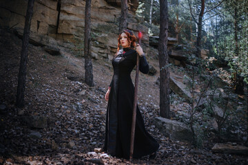 Evil witch with a staff in the forest. Slavic witch creates red magic. A young woman in a black dress and a crown embroidered with beads. - obrazy, fototapety, plakaty