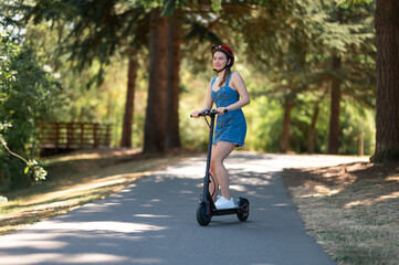 An environmentally friendly form of transport - an electric scooter. A young girl in a protective helmet and denim dress rides in the park. Courier, outdoor activities, sports.