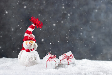 Funny Christmas snowman in knitted striped hat with pompom and gift boxes on snow. Merry Christmas and Happy New Year greeting card with copy space.