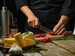A chef in a black uniform is cutting tomatoes on a cutting board. Cheese, garlic, spices, rosemary sprigs. Cooking salad, sauce, pizza, focaccia. Close-up. Wooden texture.