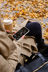Girl with a smartphone and a cup of coffee sits on a bench in an autumn park. A woman is sending a...