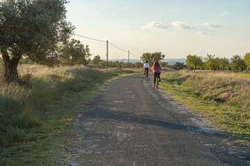 Two young girls seen from behind, riding bikes at sunset on a country road.