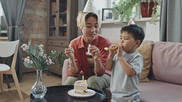 Medium slowmo shot of Asian mother and 3-year-old son in Birthday hats blowing noisemakers celebrating Birthday together at home