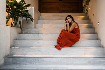 beautiful Hispanic woman in a terracotta dress sitting on the entrance stairs of a building with her hands on her face