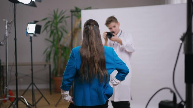 Back view teenage brunette girl posing as blurred Caucasian boy in doctor gown taking photos. Talented teenager choosing between photographer and medical occupation. Lifestyle concept
