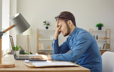 Tired worker having problem. Man makes dumb mistake or has difficulty with digital file. Side view of upset stressed young guy facepalming sitting at working desk with laptop computer in home office - obrazy, fototapety, plakaty