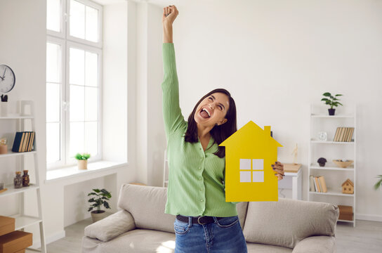 Happy Single Young Woman Who Finally Has Enough Money To Buy New Home Or Pay Off Mortgage Standing In Living Room, Holding Symbolic Paper House, Raising Hand Up And Shouting YES. Real Estate Concept