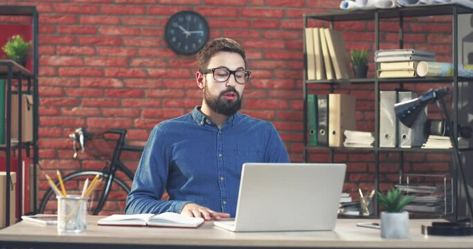 Young Caucasian businessman sits in modern office and talks to colleagues via video chat using laptop. Concept of remote work.