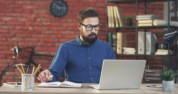 Young Caucasian businessman with beard and glasses sits in modern office and talks to colleagues on video call looking at laptop.
