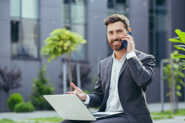 Successful male businessman reports good news by phone, working with laptop at lunchtime near office sitting on bench