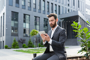 Serious male freelancer in a business suit watching an online course uses a tablet, sitting in a public place on a bench