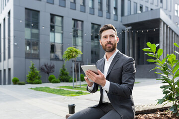 Serious male freelancer in a business suit watching an online course uses a tablet, sitting in a public place on a bench
