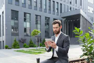 Serious male freelancer in a business suit watching an online course uses a tablet, sitting in a...
