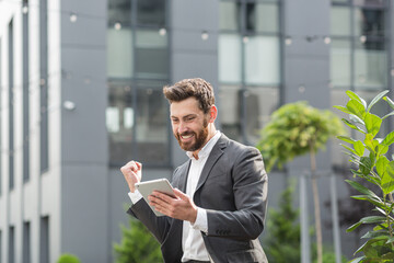 Successful male businessman got good win news reads them from a tablet computer, rejoices and...