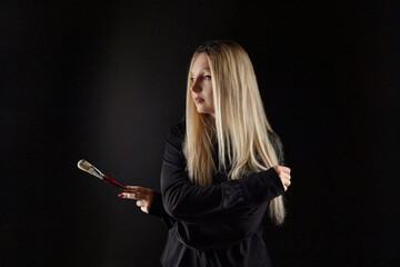 young blonde artist poses with a brush in her hand. photo shoot on a black background in the studio
