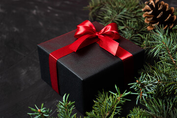 Brutal gift in black packaging with a red ribbon and a bow on a dark background with pine branches and cones