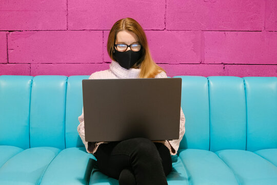 Young attractive woman working on laptop in modern coworking space with bright magenta walls and blue sofas and drinking cocoa. Elerning or freelance concept.