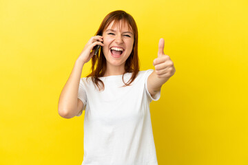 Redhead girl isolated on yellow background using mobile phone while doing thumbs up