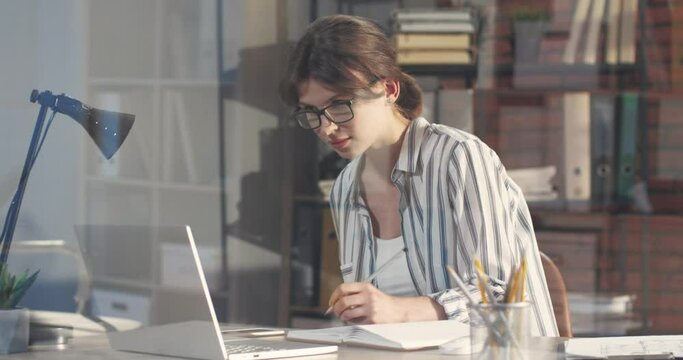 Young beautiful business woman left-handed focused working in office on laptop and making pencil notes in notebook.