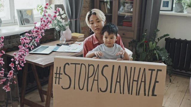 Slowmo shot of young Asian woman and her 3-year-old son posing for camera with cardboard Stop Asian Hate sign at home interior
