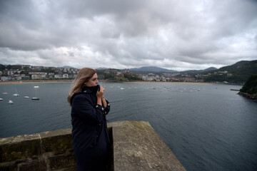 Una mujer rubia con abrigo negra mirando hacia el mar con el viento de cara y el cielo nublado