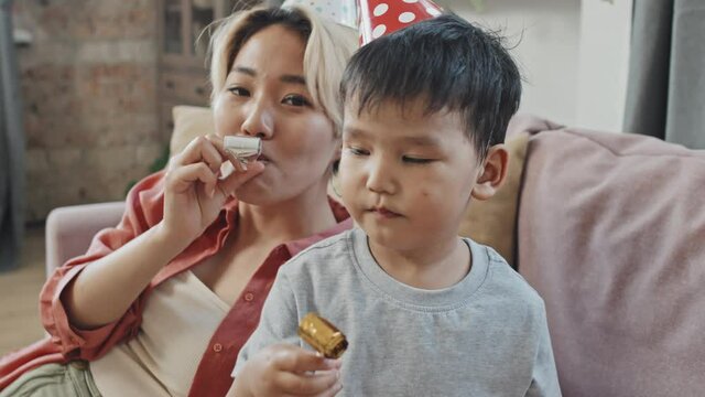 Medium close-up with slowmo of cheerful Asian mother and little son wearing funny Birthday hats blowing party horns at camera celebrating Birthday at home