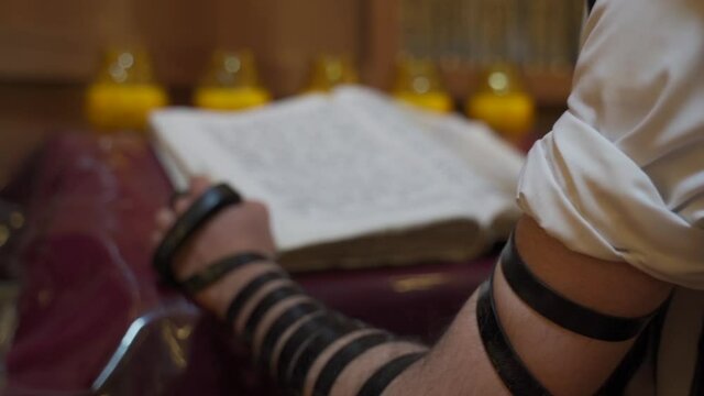 A Jew prays in the synagogue. A man stands near the presidium and reads a book. Back view