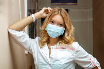 a young tired woman doctor in a mask and a white coat is leaning against the wall in the corridor of the hospital
