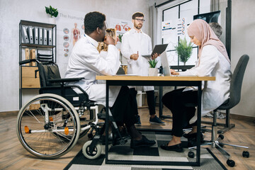 Fototapeta premium African doctor who using wheelchair sitting with his multiracial colleagues at table and listening caucasian lecturer during meeting. Medical seminar of competent specialists.