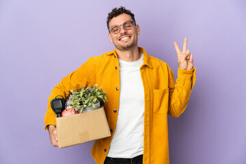 Young caucasian man making a move while picking up a box full of things isolated on purple...