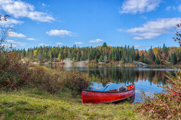 A red canoe pulled up on shore at a lake with the leaves on the trees in the background changing color in fall. 