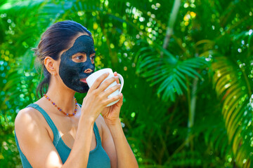 Happy woman with charcoal facemask drinks tea