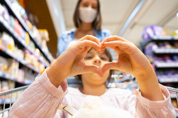 Obraz premium Pretty young girl making heart sign with her hands while sitting in the shopping cart and her mother standing behind her in a medical mask, family shopping concept.