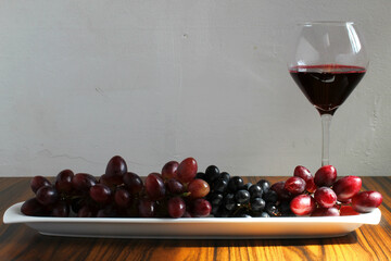 Plate with grapes and glass of wine. Front view. Wooden table and rustic white wall. Side light illuminating half of the photo