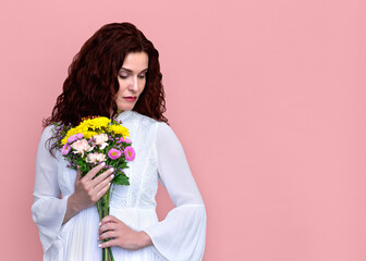 Woman Holding Flowers to Chest Looking Down on Pink Background. Portrait of sad pensive woman in white dress holding bouquet of flowers. Copy room.